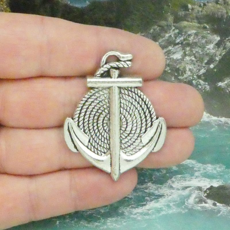 silver anchor charm with two flukes and a coiled rope rests across four fingers on a coastal background